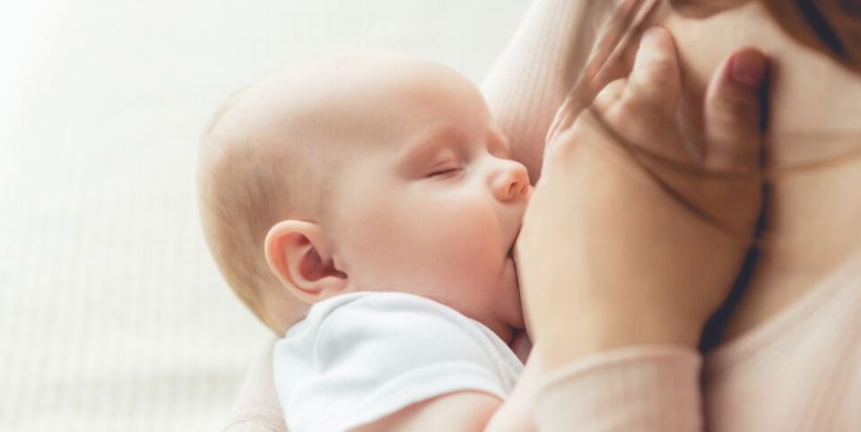 Stillendes Baby in Nahaufnahme, das an der Brust seiner Mutter trinkt. Der Hintergrund ist weich und lichtdurchflutet, mit einem sanften Fokus auf das Baby.