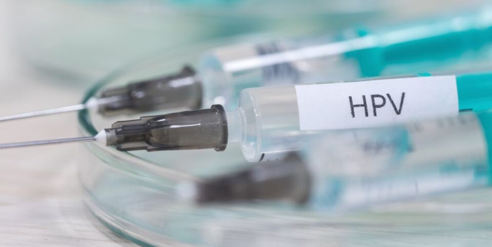 HPV-Impfstoff in einer Spritze mit Nadel, platziert auf einer gläsernen Unterlage. Unscharfer Hintergrund mit weiteren medizinischen Instrumenten.