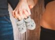 Hände eines Paares halten graue Babystrümpfe, während der Babybauch der Frau sichtbar ist – Fokus auf Vorfreude und Schwangerschaft.