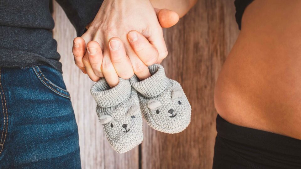 Hände eines Paares halten graue Babystrümpfe, während der Babybauch der Frau sichtbar ist – Fokus auf Vorfreude und Schwangerschaft.