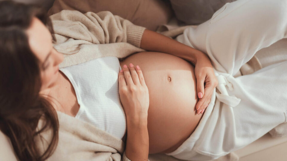 Careless lady lying and holding her hands on pregnant belly Schwangere Frau formt mit ihren Händen ein Herz über ihrem Babybauch – Symbol für Liebe und Vorfreude auf die Geburt.
