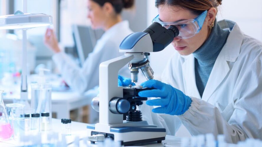 Wissenschaftlerin im Labor untersucht eine Probe unter einem Mikroskop, umgeben von Laborausrüstung.