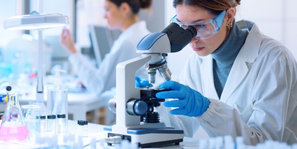 Wissenschaftlerin im Labor untersucht eine Probe unter einem Mikroskop, umgeben von Laborausrüstung.