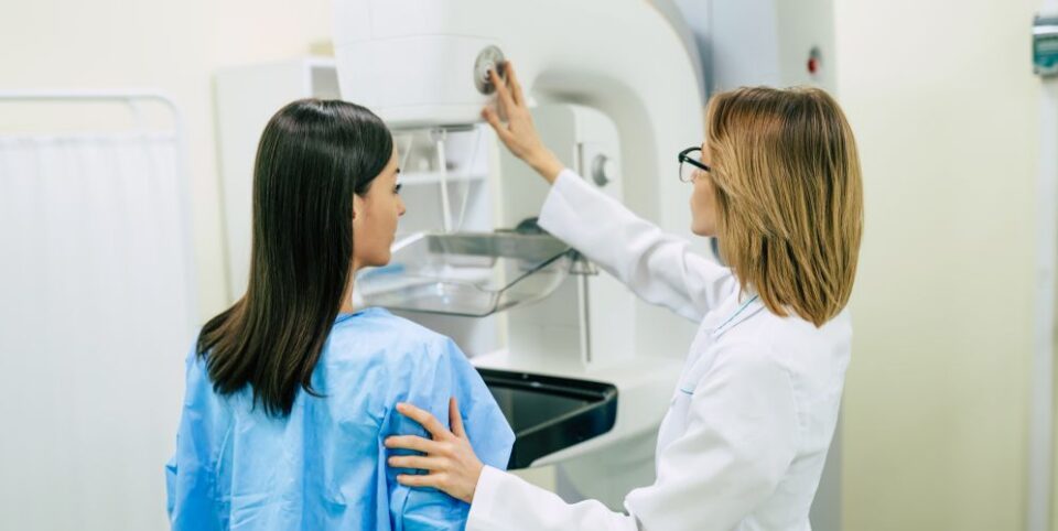Ärztin begleitet eine Patientin in einem blauen Kittel bei einer Mammographie-Untersuchung an einem modernen Gerät – Fokus auf Brustkrebsvorsorge