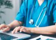Medizinisches Fachpersonal in blauer Arbeitskleidung tippt auf einem Laptop in einer Praxis oder Klinik – Fokus auf digitale Gesundheitsdokumentation.