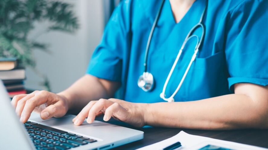 Medizinisches Fachpersonal in blauer Arbeitskleidung tippt auf einem Laptop in einer Praxis oder Klinik – Fokus auf digitale Gesundheitsdokumentation.