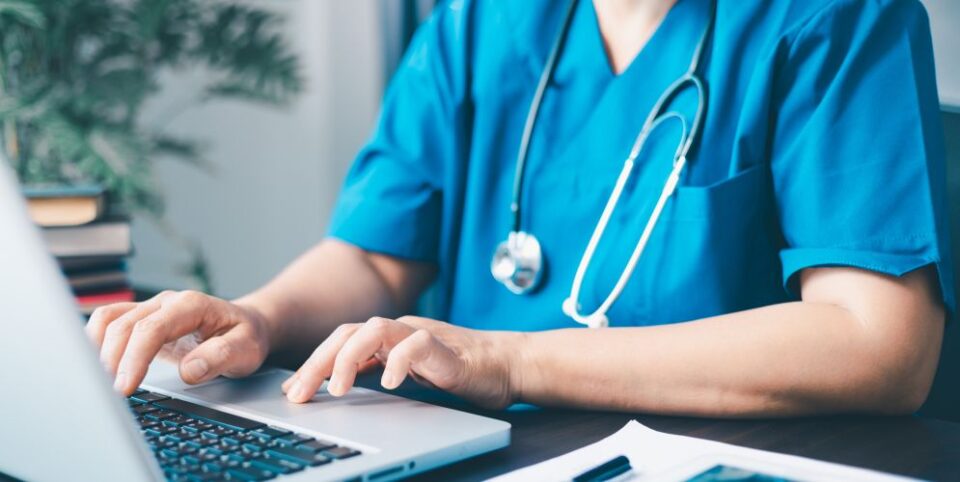 Medizinisches Fachpersonal in blauer Arbeitskleidung tippt auf einem Laptop in einer Praxis oder Klinik – Fokus auf digitale Gesundheitsdokumentation.