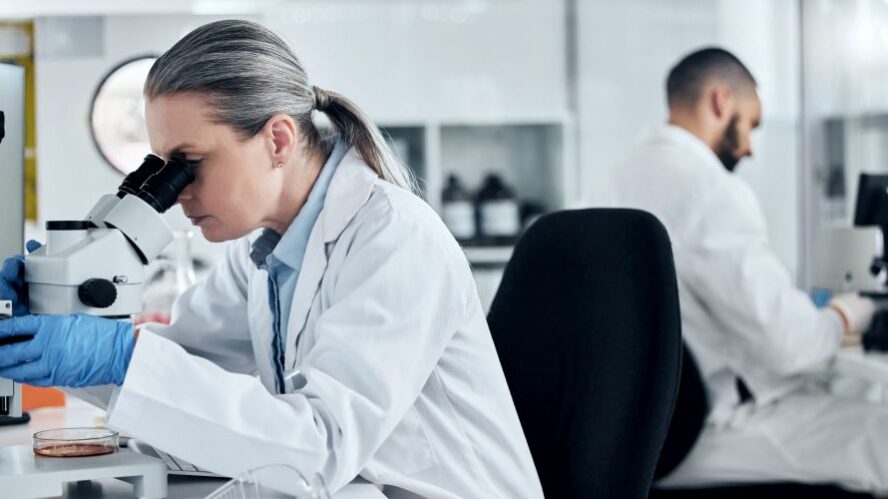 Frau im Labor untersucht eine Probe unter einem Mikroskop, während ein weiterer Wissenschaftler im Hintergrund arbeitet – Fokus auf Forschung und Laboranalysen.