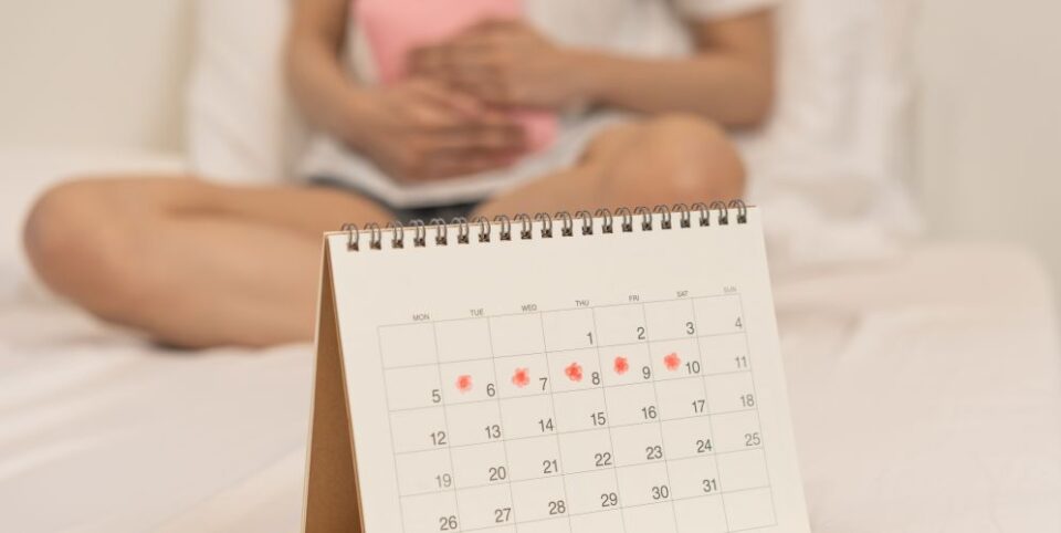 Tischkalender mit markierten Menstruationstagen im Vordergrund, während im Hintergrund eine Frau mit einer Wärmflasche ihren Bauch hält – Fokus auf Menstruationszyklus und Schmerzen.