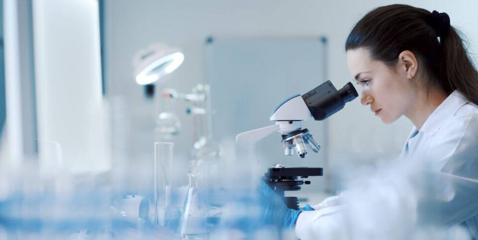 Wissenschaftlerin im Labor untersucht eine Probe unter einem Mikroskop, umgeben von Laborgeräten – Fokus auf Forschung und Analyse.