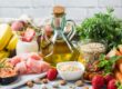 Frische Lebensmittel wie Obst, Gemüse, Olivenöl, Haferflocken, Fisch und Hähnchen arrangiert auf einem Tisch – Fokus auf gesunde und ausgewogene Ernährung.