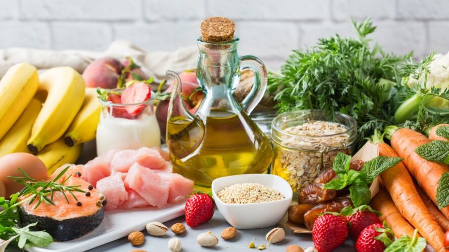 Frische Lebensmittel wie Obst, Gemüse, Olivenöl, Haferflocken, Fisch und Hähnchen arrangiert auf einem Tisch – Fokus auf gesunde und ausgewogene Ernährung.