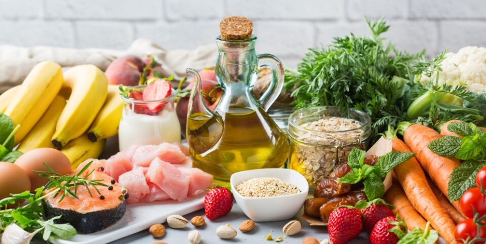 Frische Lebensmittel wie Obst, Gemüse, Olivenöl, Haferflocken, Fisch und Hähnchen arrangiert auf einem Tisch – Fokus auf gesunde und ausgewogene Ernährung.