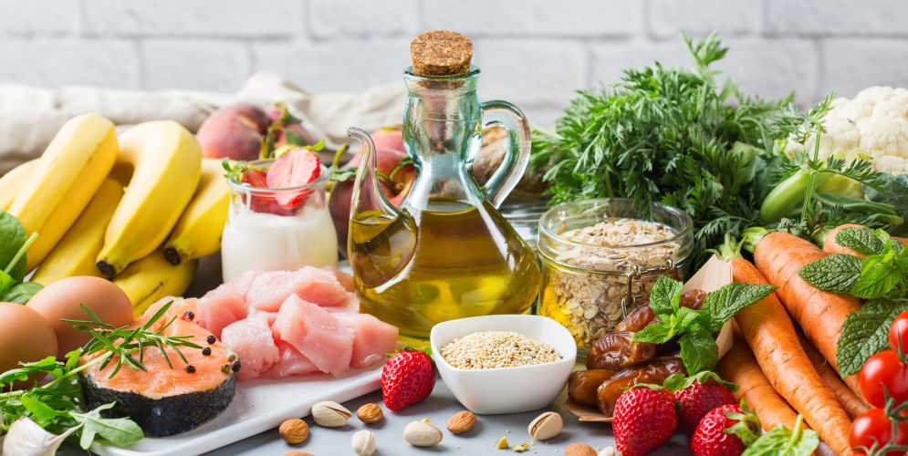 Frische Lebensmittel wie Obst, Gemüse, Olivenöl, Haferflocken, Fisch und Hähnchen arrangiert auf einem Tisch – Fokus auf gesunde und ausgewogene Ernährung.