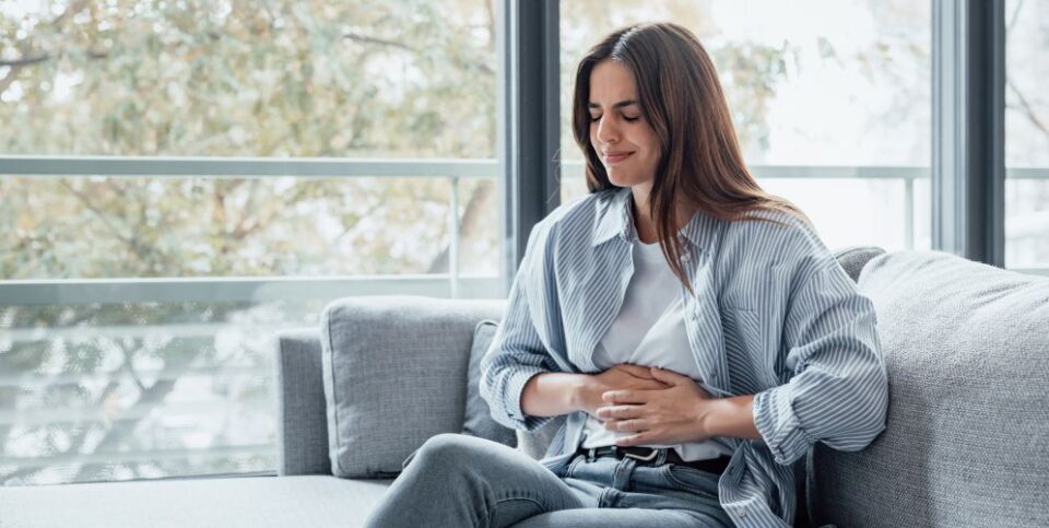 Junge Frau sitzt auf einer Couch und hält sich mit schmerzverzerrtem Gesicht den Bauch – Fokus auf Magenbeschwerden oder Bauchschmerzen.