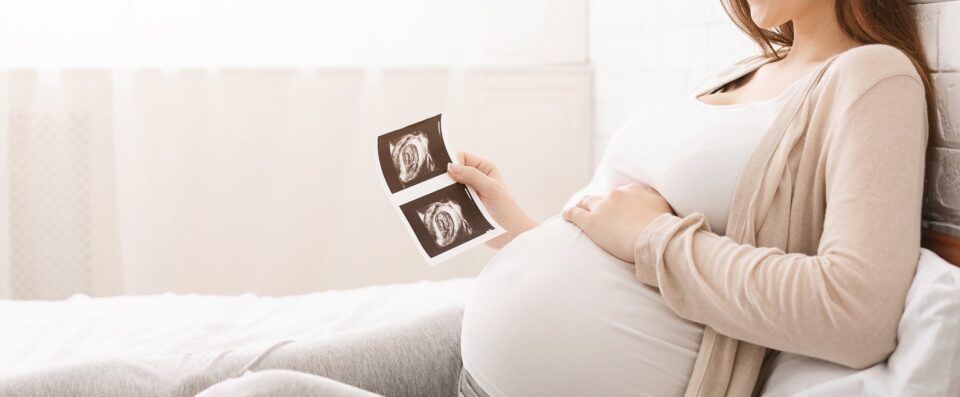 Schwangere Frau hält Ultraschallbilder in der Hand, während sie auf einem Bett sitzt. Sanfter, heller Hintergrund mit warmen Farbtönen.