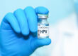 Hand in blauem Handschuh hält HPV-Impfstofffläschchen