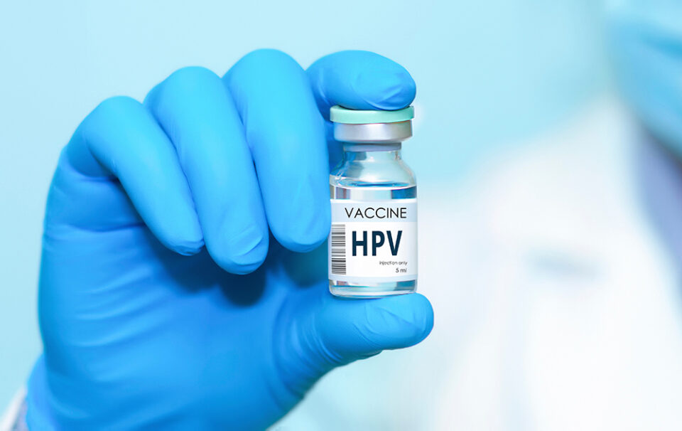Hand in blauem Handschuh hält HPV-Impfstofffläschchen